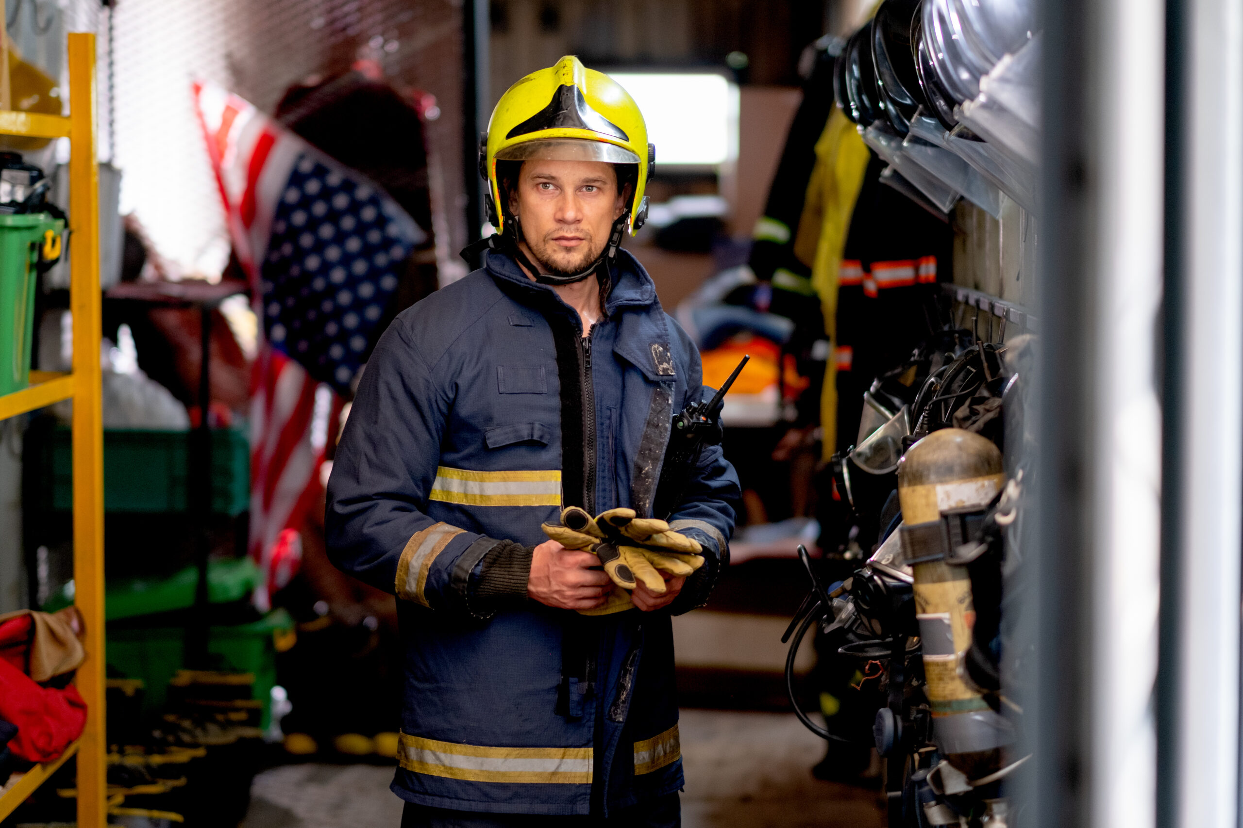 Fireman or firefighter man stand and hold the glove and look at camera also stay in room with many tools or equipment for extinguish fire in other place and support safety for people.
