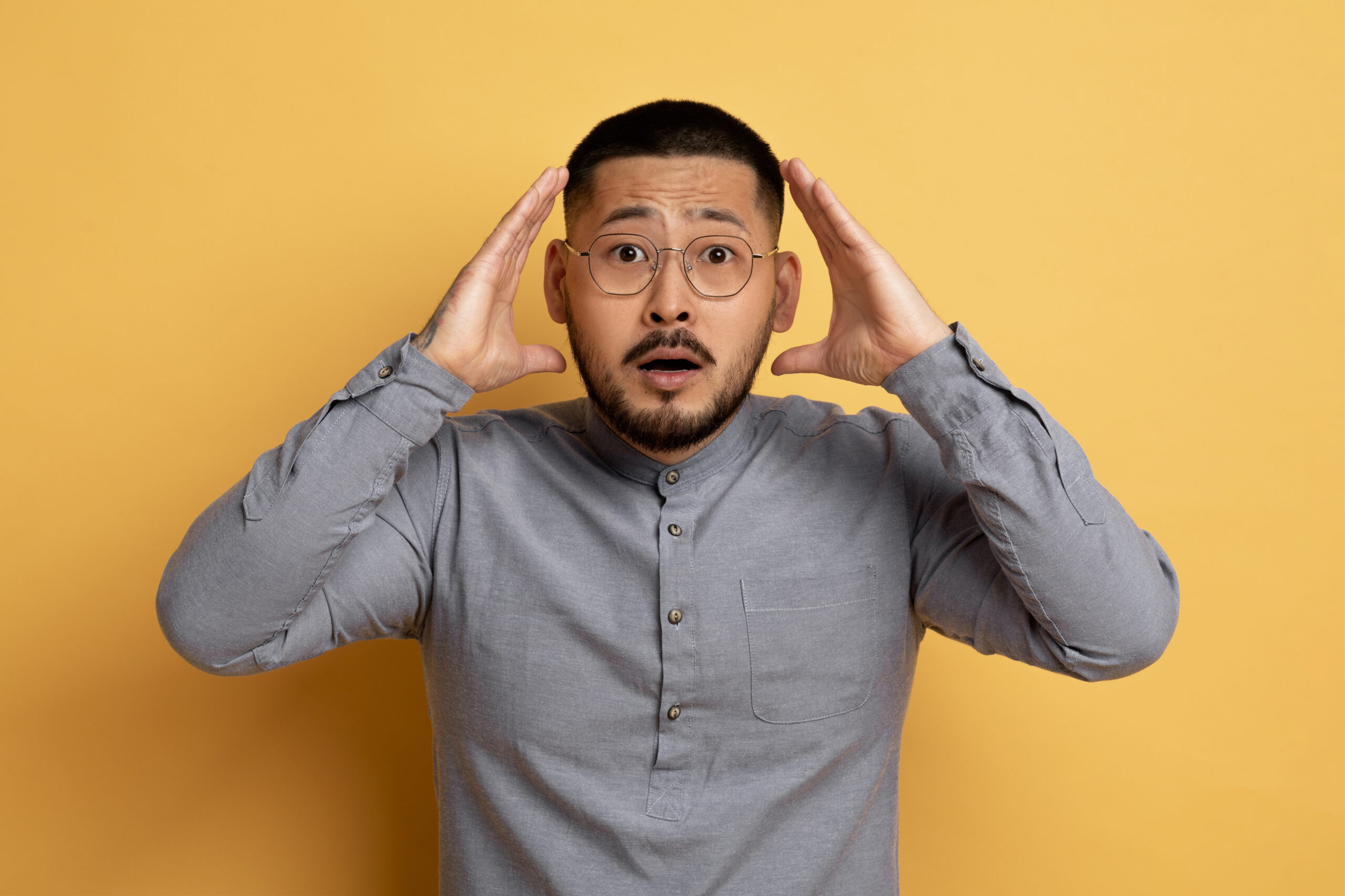 Portrait Of Shocked Young Asian Man Touching Head And Looking At Camera, Terrified Millennial Male In Eyeglasses Emotionally Reacting To Bad News, Standing Over Yellow Studio Background, Free Space