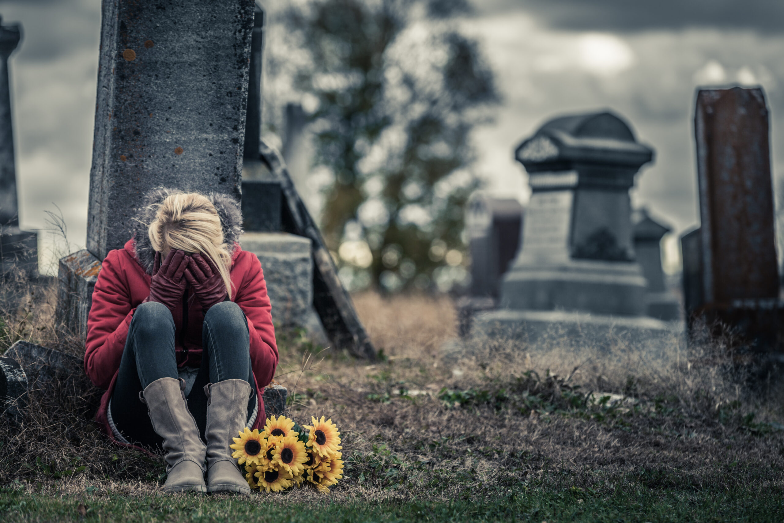 Lonely Crying Young Woman in Mourning with Sunflowers in front of a Gravestone in a Cemetery