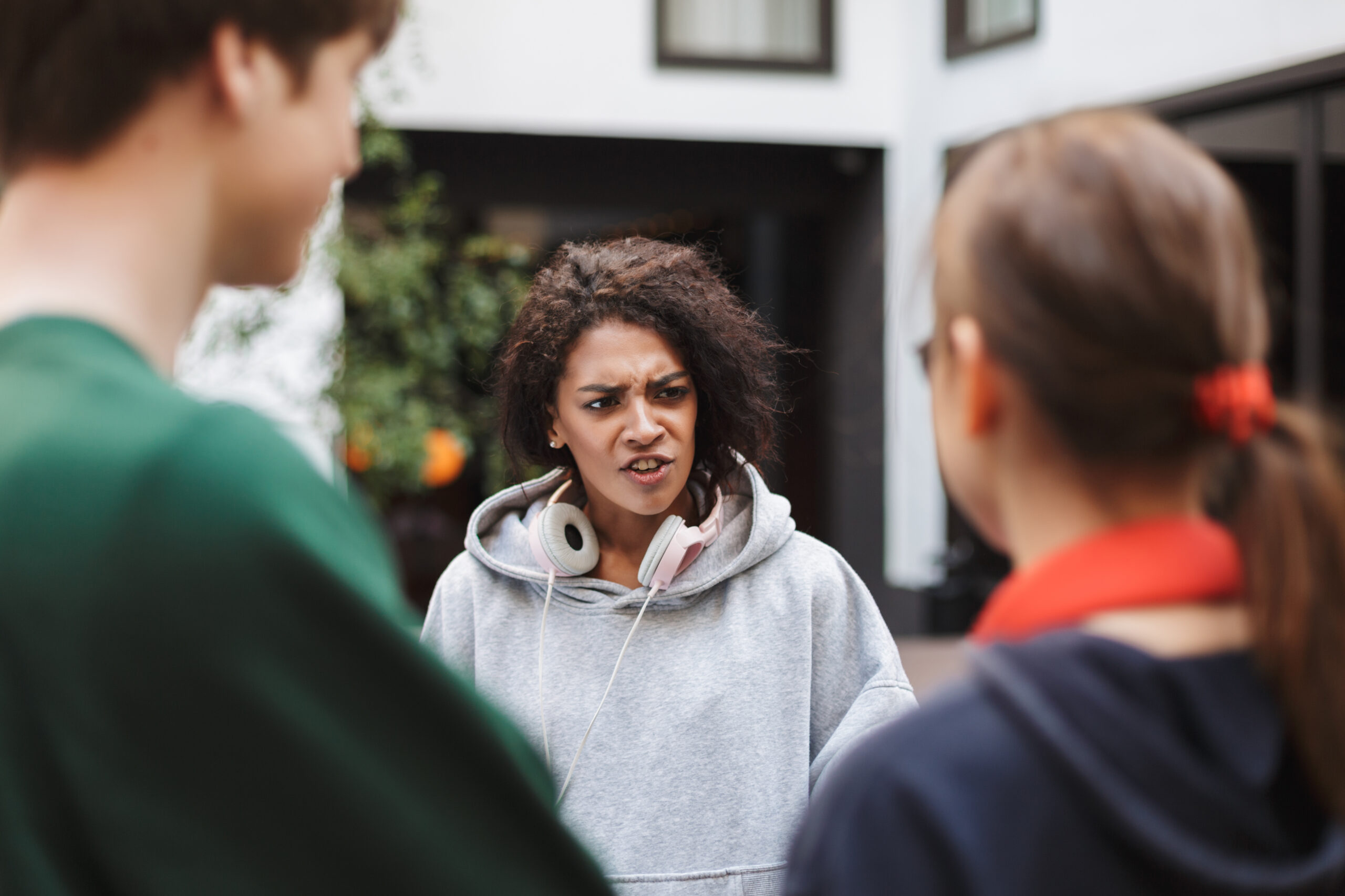 Young emotional lady with dark curly hair in headphones standing and discussion study with another students in courtyard of university