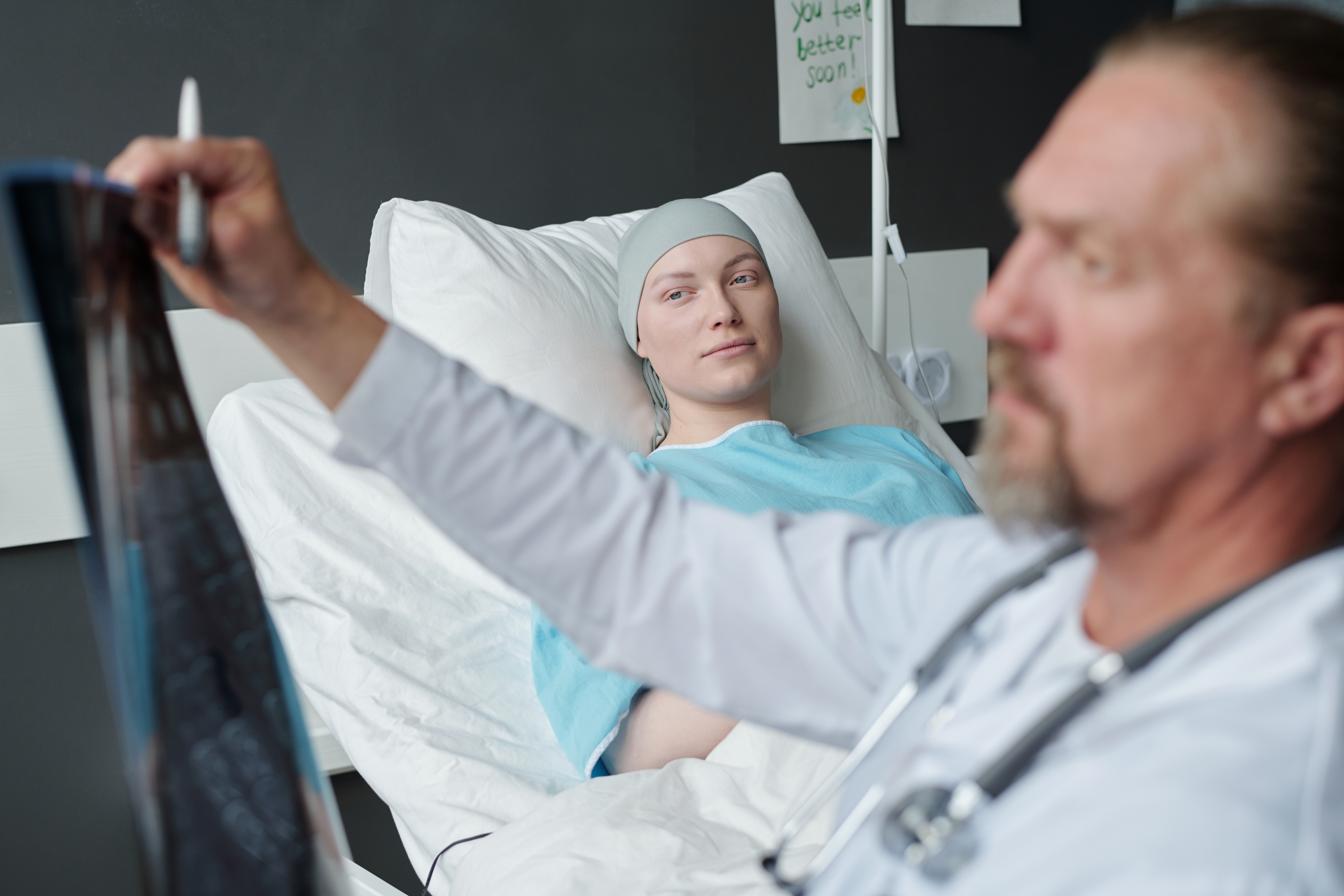 Young woman with brain cancer lying in bed and looking at oncologist analyzing x-ray image with results of MRI scan in hospital ward