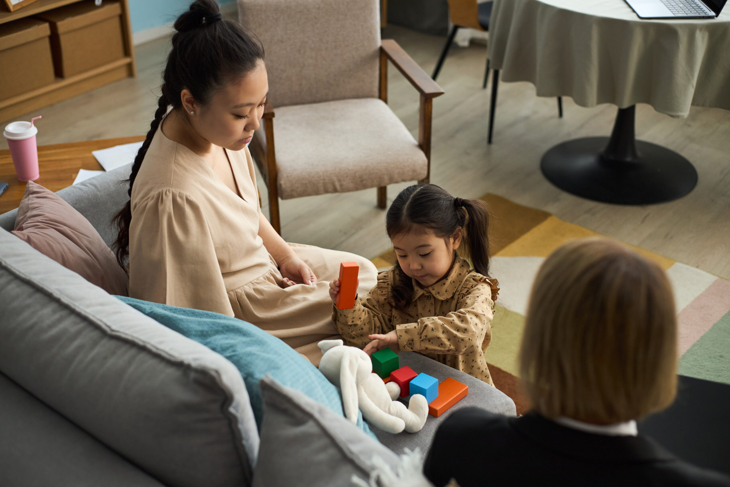 Woman sitting on couch, observing child playing with blocks, young mother focusing on activity in cozy living room environment