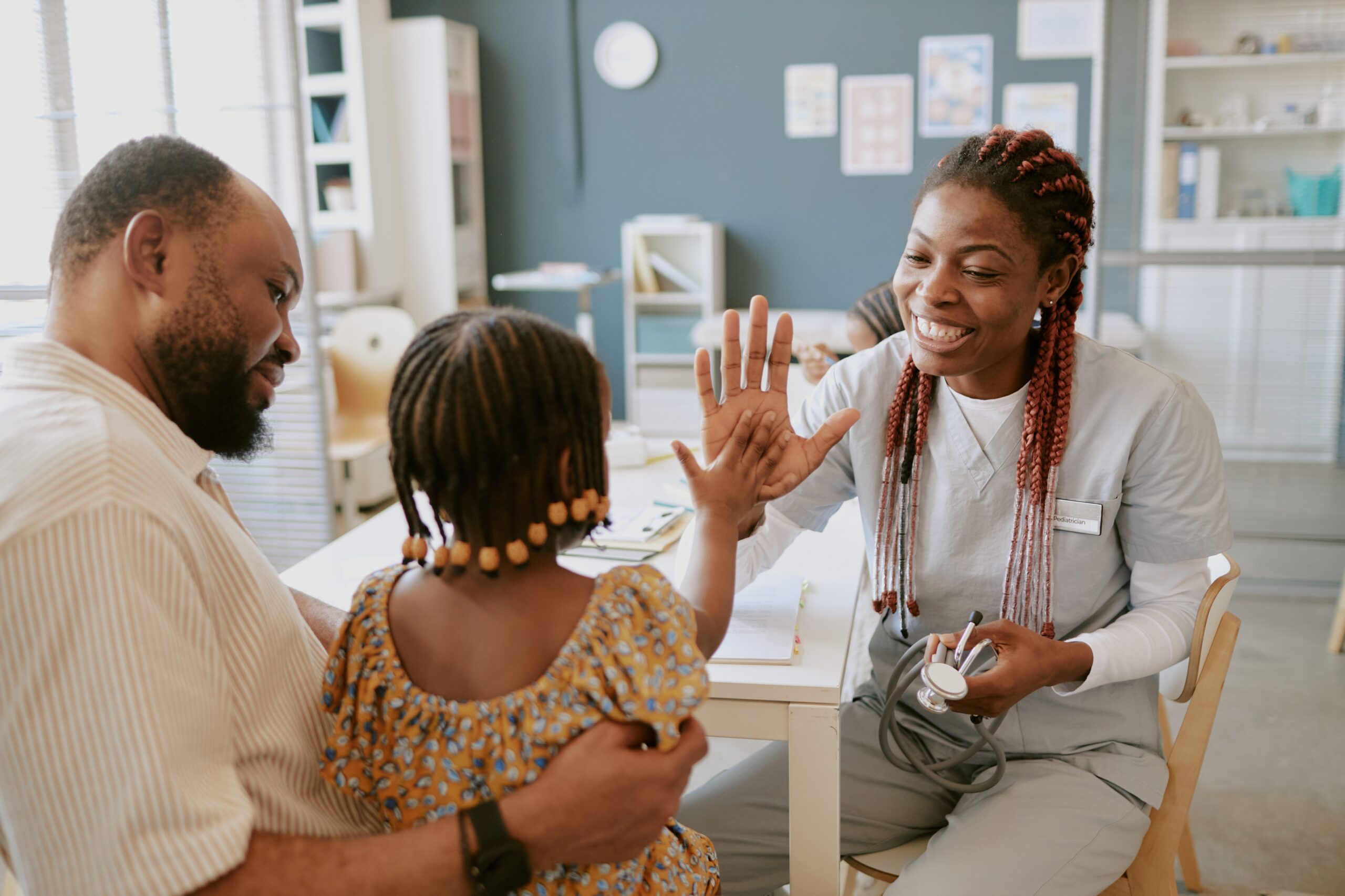 Healthcare worker smiling and greeting young child with an arm around their father in a contemporary medical office with blue walls